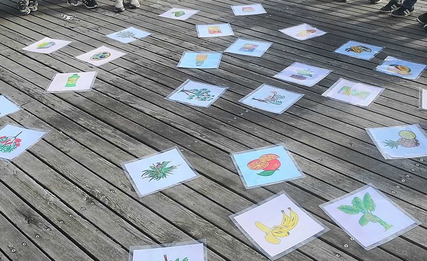 Auf Holzplanken liegen laminierte Zeichnungen von Lebensmitteln wie Bananen oder Ananas, im Hintergrund sieht man angeschnitten eine Gruppe von Jugendlichen